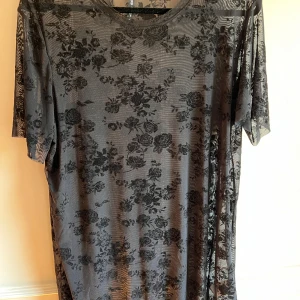 Svart blommig mesh-topp från Cropp - Säljer en svart blommig mesh-topp från Cropp. Toppen har en transparent design med blommönster och korta ärmar. Perfekt för att lägga till en edgy touch till din outfit.