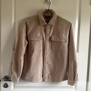 Beige manchester overshirt från Tommy Hilfiger - Säljer en snygg beige overshirt i manchester från Tommy Hilfiger. Skjortan har långa ärmar och två bröstfickor med knappar. Perfekt för lager-på-lager-stil. Klassisk design med en modern touch.