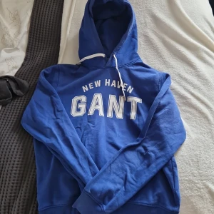 Blå hoodie från GANT - Säljer en snygg blå hoodie från GANT med texten 'NEW HAVEN GANT' tryckt på framsidan. Tröjan har en klassisk känguruficka och justerbar huva med vita snören. Perfekt för en avslappnad stil.