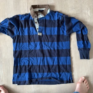 Blå och svart randig pikétröja från Gant - Säljer en stilren blå och svart randig pikétröja från Gant. Tröjan har en brun krage i manchester och knappar framtill. Perfekt för en avslappnad men ändå snygg look.