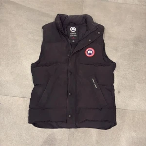 Svart dunväst från Canada Goose - Säljer en stilren svart dunväst från Canada Goose med ett ikoniskt märke på bröstet. Västen har dragkedja och knappar framtill för extra skydd mot kyla. Perfekt för lager-på-lager under kyliga dagar.
