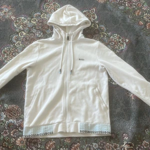 Vit hoodie från Boss - Säljer en stilren vit hoodie från Boss med dragkedja och huva. Tröjan har långa ärmar och ett elastiskt band med BOSS-logga vid nederkanten. Perfekt för en avslappnad stil.