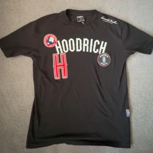 Svart t-shirt från Hoodrich - Snygg svart t-shirt från Hoodrich med vita och röda detaljer. Den har korta ärmar och flera coola patches på framsidan. Perfekt för en avslappnad stil. Slutsåld på marknaden men hos mig får ni den med ett utmärkt pris och utmärkt skick.