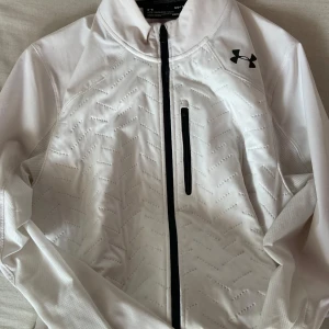 Vit träningsjacka från Under Armour - Säljer en vit träningsjacka från Under Armour med dragkedja och en bröstficka. Jackan har långärmade ärmar och är tillverkad i syntetmaterial. Perfekt för kyligare dagar med ColdGear-teknologi.