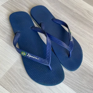 Blå flip-flops från Ipanema - Säljer ett par klassiska blå flip-flops från Ipanema med brasiliansk flagga på remmen. Perfekta för stranden eller sommardagar. Lätta och bekväma med platt sula och enkel design.