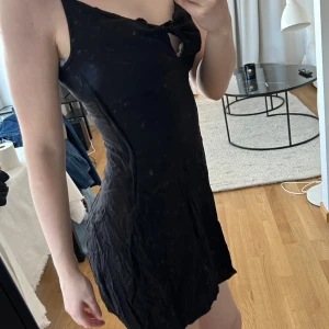 Svart kort klänning med knyt fram - Säljer en svart kort klänning med smala axelband och knytning framtill vid bysten. Klänningen har en figurnära passform och är perfekt för dig som gillar en enkel men snygg stil. Materialet känns lätt och smidigt.
