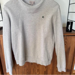 Grå tröja från Morris - Säljer en stilren grå tröja från Morris med ett litet emblem på bröstet. Tröjan har långa ärmar och en rund halsringning.