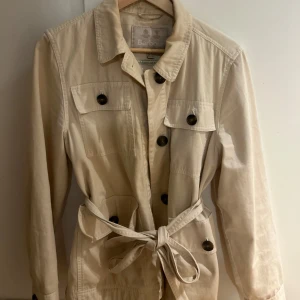 Barbour coat, limited edition  - Säljer en stilren beige jacka från Barbour med knappar och knytbälte i midjan. Jackan har långa ärmar och två bröstfickor med knappar. Perfekt för en klassisk look. köpt på Johnells med Nypris 5560kr, endast använd en höst
