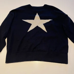 Mörkblå stickad stjärntröja  från Gina Tricot - Säljer en mörkblå stickad tröja från Gina Tricot med en stor vit stjärna på framsidan. Tröjan har lång ärm och rund halsringning. .