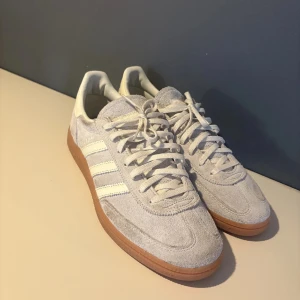 Adidas Spezial sneakers i ljus mocka - Säljer ett par Adidas Spezial sneakers i ljusgrå mocka med vita detaljer och klassiska tre ränder på sidan. Skorna har snörning och en brun gummisula som ger en retro känsla. Perfekta för dig som gillar stilrena och tidlösa sneakers.