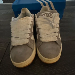 Adidas Campus grå sneakers - Grå Adidas Campus sneakers med vita ränder och vit sula. Skorna har klassisk låg modell, rund tå och breda vita skosnören. Ovandelen är i mocka och yttersulan är i gummi. Adidas logga syns på häl och plös. Använt kanske 1 eller 2 gånger ute