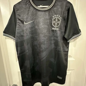 Svart Brasil Nike fotbollströja - Snygg svart Brasil fotbollströja från Nike med diskret camouflagemönster. Tröjan har korta ärmar, broderat CBF-märke och Nike-logga på bröstet samt vita detaljer på krage och ärmslut. Tillverkad i lätt och ventilerande material, perfekt för match eller träning.