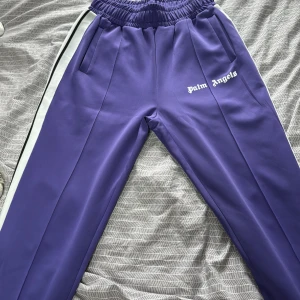 Palm Angels lila mjukisbyxor M - Palm Angels mjukisbyxor i lila med vita och svarta sidoränder. Byxorna har elastisk midja, raka ben och logga broderad på vänster sida. Tillverkade i polyester för en sportig vibe. Perfekta för en chill och trendig streetwear-look. Köpta i London 2020