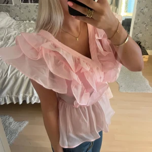 Rosa volangblus med v-ringning - Supersöt ljusrosa blus med stora volanger och v-ringning. Blusen har knappar framtill och är tillverkad i ett tunt, lätt och transparent material som ger en fluffig känsla. Perfekt för dig som vill sticka ut med en romantisk och feminin look.