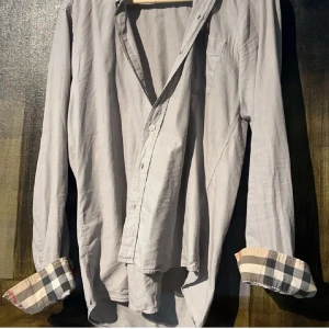 Grå skjorta från Burberry med rutiga detaljer - Snygg grå skjorta från Burberry med klassiska rutiga detaljer på insidan av kragen och ärmsluten. Perfekt för dig som gillar stilrena plagg och perfekt inför sommaren🥂