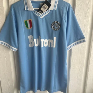 Napoli Maradona 10 retro fotbollströja - Retroinspirerad ljusblå Napoli fotbollströja med Maradona 10 på ryggen. Klassisk vit krage med blå detaljer, broderad klubbmärke och italienskt mästerskapsmärke på bröstet. Tillverkad i lätt och ventilerande material, perfekt för fans av italiensk fotboll.