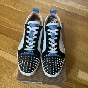 Christian Louboutin/loubs - Snygga sneakers från Christian Louboutin med vit, svart och blå färg. Skorna har coola silverfärgade nitar på tån, vit sula och den klassiska röda undersidan. Materialmix av skinn och mocka, med vita snören och blå detaljer på plösen och hälen. Skorna är alltid rengjorda efter användning. De sitter aningen stort