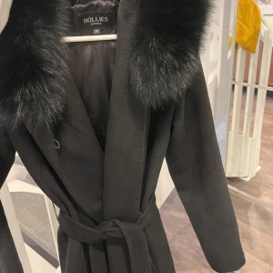 Svart kappa från Hollies - Snygg svart kappa från Hollies med stor krage och knytskärp i midjan. Klassisk och stilren modell med långa ärmar och mjukt ullmaterial. 100% ull. 