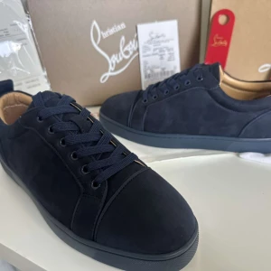 Mörkblå sneakers från Christian Louboutin - Snygga mörkblå sneakers från Christian Louboutin i mocka med klassisk röd sula och broderad logga på hälen. Skorna har rund tå, platt sula och matchande mörkblå snörning. Insidan är fodrad i beige läder för extra komfort och lyxig känsla.