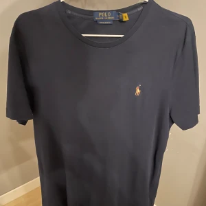 Marinblå t-shirt från Polo Ralph Lauren - Snygg mörkblå t-shirt från Polo Ralph Lauren med klassisk passform och broderad logga på bröstet. Tillverkad i mjuk bomull och har rund halsringning samt korta ärmar. Perfekt för en clean och stilren look. Knappast användt den då den är lite för stor för mig. Så den är i toppskick. Bara att höra av sig vid minsta fundering!