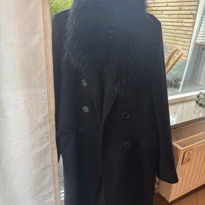 Svart kappa med fuskpäls från Zara Woman - Stilren svart kappa från Zara Woman med bred krage i fuskpäls och dubbla knapprader. Tillverkad i ullblandning från Manteco, vilket ger en lyxig känsla. Perfekt för dig som vill ha en klassisk och trendig look under höst och vinter.