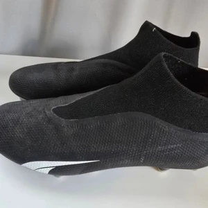 Svarta Puma Ultra fotbollsskor - Säljer ett par svarta Puma Ultra fotbollsskor med vit logga på sidan. Skorna har strumpliknande skaft och ovandel i syntetmaterial för en tight passform. Sulan har fasta dobbar för gräsplan. Riktigt clean och modern look för dig som vill sticka ut på planen.