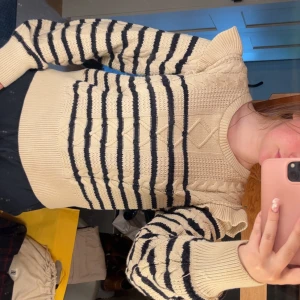 Beige och svart randig stickad tröja - Superfin stickad tröja i beige med svarta ränder och snygga kabelstickade detaljer framtill. Tröjan har volang vid axlarna och långa ärmar med breda muddar. Perfekt för dig som gillar en trendig och mysig look.