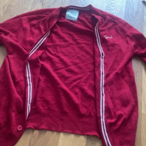 Röd kofta i lammull från Gant XL - Röd kofta från Gant i 100% lammull med vita ränder längs insidan av knappslån. Klassisk design med broderad Gant-logga på bröstet och knapp nertill. Perfekt för lager-på-lager och riktigt skön att ha på sig under kyliga dagar.