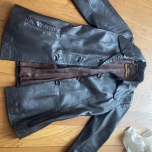 Brun skinnjacka från Torsten Albertssons - Snygg brun skinnjacka från Torsten Albertssons med klassisk krage och knäppning framtill. Jackan har raka linjer, två fickor och är helfodrad med brunt innerfoder. Perfekt för dig som gillar retrovibbar och tidlös stil.