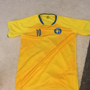 Sverige fotbollströja gul nr 10 H&M - Gul fotbollströja från H&M med blå detaljer, nummer 10 på både fram- och baksida samt texten 'Sverige' på ryggen. Tröjan har korta ärmar, blå krage och blått tryck. Perfekt för dig som vill heja på Sverige med stil.