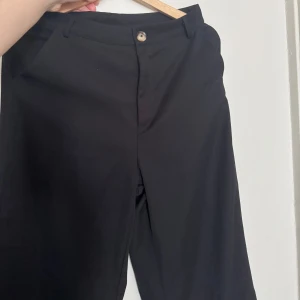 Svarta kostymbyxor med rak passform - Stilrena svarta kostymbyxor med klassisk rak passform och midja med knappstängning. Byxorna har hällor för bälte och sidfickor. Perfekta för en clean och dressad look. Materialet känns mjukt och lätt, troligtvis polyester eller bomullsmix.