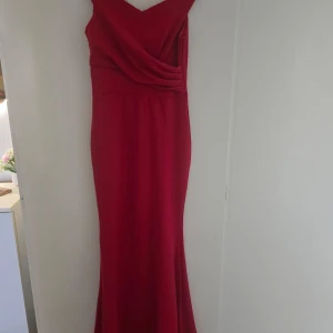 Röd långklänning med omlott-detalj - Snygg röd långklänning med omlott-drapering över bysten och figurnära passform. Klänningen har ärmlös design och en elegant v-ringning. Perfekt för bal eller festliga tillfällen. Materialet känns mjukt och följsamt.