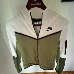 Nike hoodie jacka i vitt och olivgrönt - Nike hoodie jacka i vitt och olivgrönt med svart logga på bröstet. Jackan har dragkedja framtill, huva och en cool färgblock-design. Ärmarna är långärmade och jackan har en ficka med dragkedja på ena ärmen. Perfekt för en sportig och avslappnad stil.