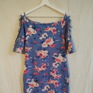 Blommig blå kortklänning med snörning - Superfin blå kortklänning med rosa och vita blommor. Klänningen har korta ärmar med snörningsdetalj längs axlarna och en figurnära passform. Perfekt för sommarens alla tillfällen när du vill känna dig extra söt och färgglad.