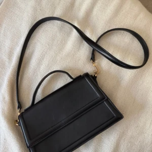 Svart axelväska från Gina Tricot - Snygg svart axelväska från Gina Tricot i stilren design. Väskan har ett avtagbart axelband, ett litet handtag och ett stort fack med dragkedja inuti. Tillverkad i slätt syntetmaterial med guldfärgade detaljer.