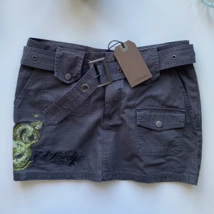ED HARDY MINIKJOL - Grå kortkjol från Ed Hardy med coolt grönt drakbroderi på sidan, stora fickor framtill och bredt bälte med metallspänne. Snygga detaljer som knappar och broderad logga. Perfekt för dig som vill sticka ut med edgy streetstyle.