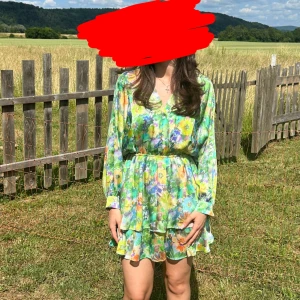 Blommig grön kortklänning med volang - Superfin kortklänning med grönt och blommigt mönster i blått, gult och lila. Klänningen har v-ringning, långa ärmar och markerad midja med volangdetaljer nertill. Perfekt för sommarens alla tillfällen!