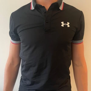 Svart pikétröja från Under Armour - Svart pikétröja från Under Armour med vit logga på bröstet. Passar perfekt till golf, tennis och tränings tröja eller bara allmänt, riktigt snyggt 