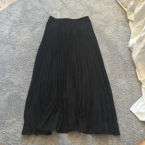 Svart plisserad långkjol - Snygg svart långkjol med plisserade veck och elastisk midja. Kjolen har ett luftigt och flowy fall som ger en stilren och trendig look. Perfekt att matcha med en crop top eller stickad tröja.