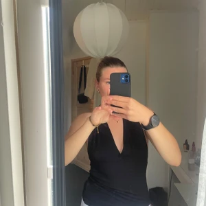 Svart halterneck topp med djup v-ringning - Säljer en svart halterneck topp med djup v-ringning och knyt i nacken. Toppen är ärmlös och har en tight passform som framhäver axlarna och ryggen. Perfekt för dig som vill ha en stilren och enkel look med lite edge.