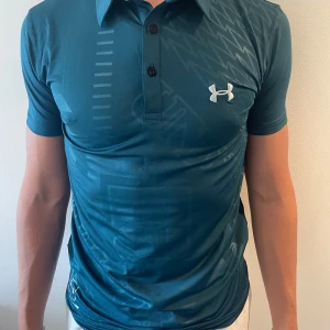 Blå pikétröja från Under Armour - Snygg blå pikétröja från Under Armour med diskret mönster och vit logga på bröstet. Passar perfekt till Golf, tennis eller om du bara vill ha snyggt