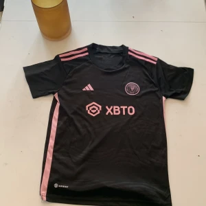 Inter Miami tröja - Säljer en snygg svart fotbollströja från Adidas med rosa detaljer. Tröjan har korta ärmar och klubbmärket för Miami på bröstet  . 💕