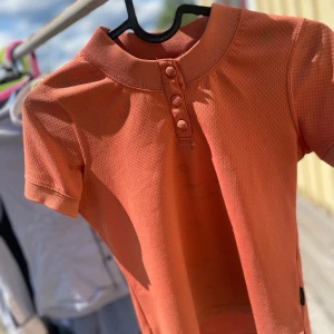 Orange kortärmad topp JH Collection - Säljer en orange kortärmad topp från JH Collection. Toppen har en ribbad hög krage och tre klädda knappar framtill. Materialet är lätt och andas, perfekt för varmare dagar. Snygg och enkel att matcha till olika outfits. ( fins en matchande kofta kontakta mig vid intresse av exempel bild )