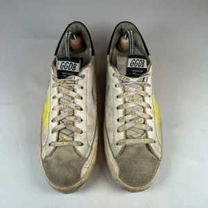 Golden Goose Superstar - Snygga sneakers från Golden Goose med vit bas, grå mockadetaljer och svart häl. Sidan pryds av en stor gul stjärna och texten 'HAVE A GOLDEN DAY' på sulan. Skorna har snörning och en cool, sliten look som sticker ut. Ingen box