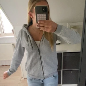 Grå hoodiejacka från Sail Racing - Grå hoodiejacka från Sail Racing med dragkedja framtill, broderad logga på bröstet och huva. Jackan har en bröstficka med dragkedja och är tillverkad i mjukt bomullsmaterial. Perfekt för chill dagar eller när du vill ha en sportig look. Tröjan har en liten fläck på framsida som troligen kan gå att få bort.