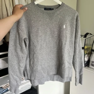 Grå sweatshirt från Polo Ralph Lauren - Klassisk grå sweatshirt från Polo Ralph Lauren med broderad vit logga på bröstet. Tröjan har rund halsringning, ribbade muddar och lång ärm. Perfekt basic att ha i garderoben, enkel att matcha och superbekväm.