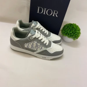 Dior sneakers i grått och vitt - Snygga Dior sneakers i vitt och grått med klassiskt Dior-monogram på sidan. Skorna har rund tå, snörning och perforerade detaljer på tån. Tillverkade i skinn och textil för en lyxig känsla. Perfekta för dig som vill sticka ut med exklusiv streetstyle.