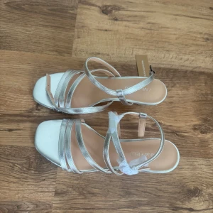 Silvriga sandaler med klack från Anna Field - Snygga sandaler i metallic silver från Anna Field med smala remmar och fyrkantig tå. Skorna har en låg klack och justerbar rem runt ankeln. Perfekta för att lyfta din outfit med en glansig touch.