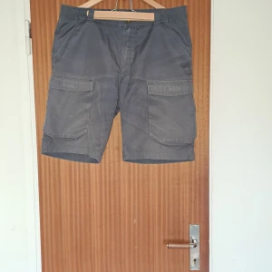 Mörkgrå cargoshorts från Everest - Snygga mörkgrå cargoshorts från Everest med flera stora fickor på sidorna och bak. Klassisk passform med bälteshällor och knappstängning. Perfekta för sommardagar när du vill ha både stil och funktion. Materialet känns slitstarkt och bekvämt.