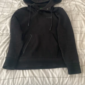 Svart hoodie från BOSS med huva - Svart hoodie från BOSS med diskret logga på bröstet, huva med snörning och stor magficka. Tröjan har långa ärmar och är tillverkad i mjuk bomullsmix, perfekt för chill dagar. Snygg och enkel design som funkar till det mesta.